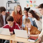 Grupo de estudantes trabalha em laptops em uma sala de aula, orientado por um professor.