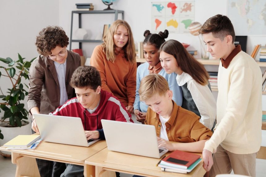 Grupo de estudantes trabalha em laptops em uma sala de aula, orientado por um professor.