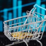 Miniatura de carrinho de compras cheia de moedas diante de um gráfico financeiro digital.